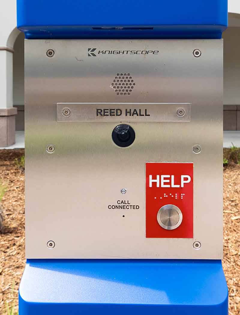 A close-up of a blue and silver emergency call box, with a bright red sticker that reads Help over a silver button. Other words on the pole says Knightscope with a company logo, Reed Hall and Call Connected. A black circle in the middle of the box indicates a security camera.