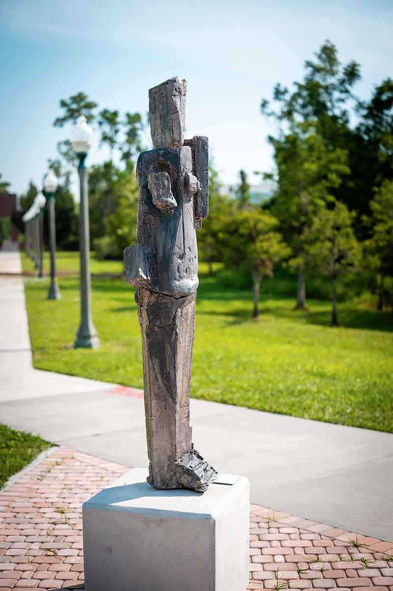 A ceramic statue of a life-sized, stylized human figure stands on a small platform with a sidewalk, lampposts, trees and grassy areas visible in the background