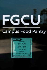 The window on a door reads FGCU Campus Food Pantry and lists the hours of operation for fall abnd spring as Monday and Friday 12-5pm, and Tuesday, Wednesday and Thursday as 1-6pm. Other words on the sign read Summer Mon thru Fri 12-5pm, and Kleist 102, by Food Forest.