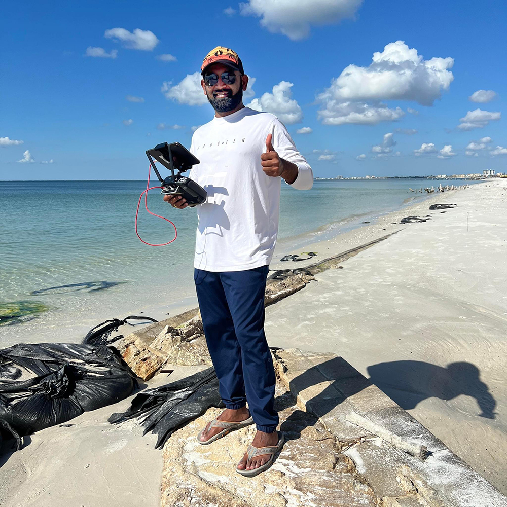 Person on a rocky beach holding a drone controller and giving a thumbs-up, with ocean waves, clouds, and black bags on the sand nearby