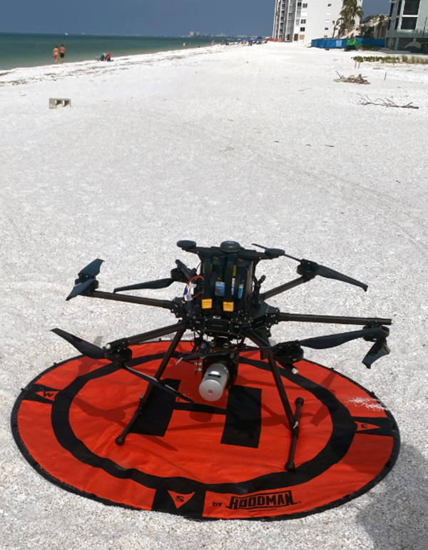 A large multi-propeller drone sits on an orange landing pad placed on a sandy beach. The ocean is visible to the left, and buildings and distant beachgoers appear in the background
