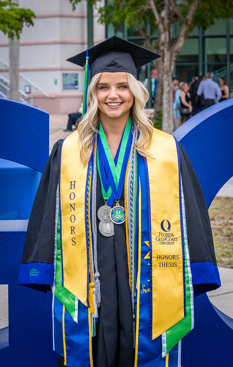 Graduate in cap and gown with honors stoles stands before blue sculpture at FGCU.