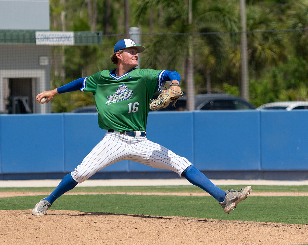 A baseball pitcher in a green jersey with the number 16 and 'FGCU' logo is captured mid-pitch on a baseball field. The pitcher is wearing white pinstriped pants, blue socks, and gray cleats. The background includes a blue fence, some parked cars, and palm trees