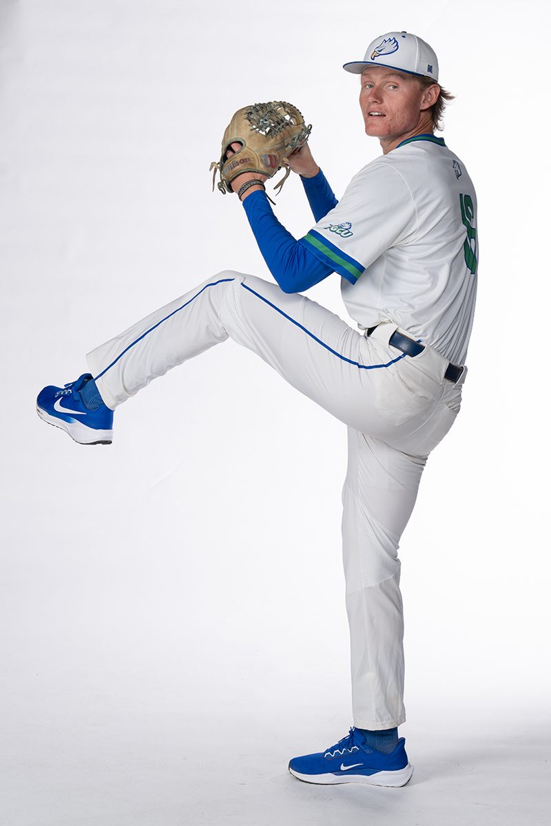 A baseball player in a white and blue uniform is captured in mid-pitching motion. The player wears a white cap with a logo, a blue undershirt, and blue cleats. The left leg is raised, and the right arm holds a glove. The background is plain white.
