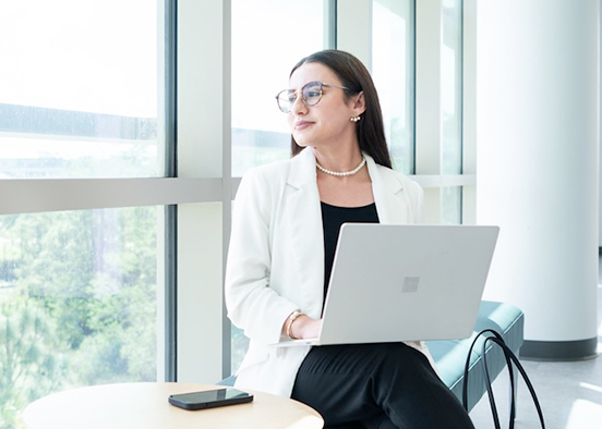 A woman is sitting on a teal bench in a modern, well-lit room with large windows. The person is wearing a white blazer, black pants, and black shoes, and is working on a laptop placed on their lap. A small round table next to the bench holds a smartphone, and a beige and black handbag rests on the floor beside the bench.