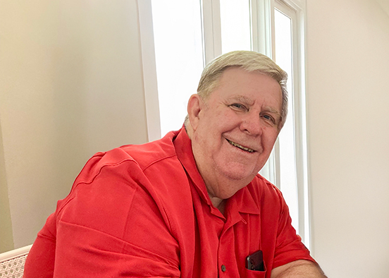 A man is sitting indoors near a window and wearing a red collared shirt and has light-colored hair. The background shows white walls and large windows allowing natural light to enter the room.