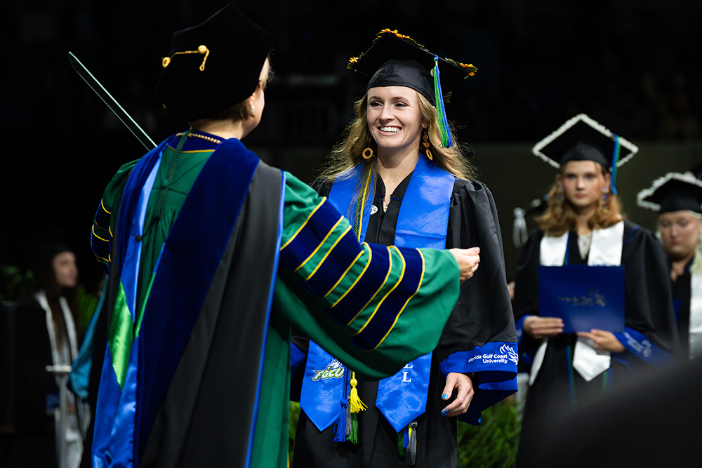 A graduation ceremony where a graduate in a black cap and gown with blue and gold tassels is receiving a diploma from an academic official dressed in green and blue regalia. Other graduates in similar attire are visible in the background, holding their diplomas