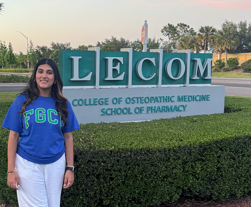 Woman stands in front of a 'LECOM College of Osteopathic Medicine School of Pharmacy' sign, wearing a blue 'FGC' shirt and white pants. Trees and a road are visible in the background.