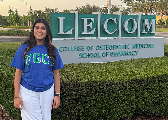 Woman stands in front of a 'LECOM College of Osteopathic Medicine School of Pharmacy' sign, wearing a blue 'FGC' shirt and white pants. Trees and a road are visible in the background.