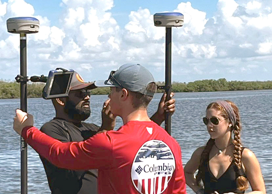 Three people stand in shallow water near a large body of water under a partly cloudy sky. Two men hold tall poles with GPS devices, and a woman stands nearby observing. They appear to be conducting a survey or fieldwork.