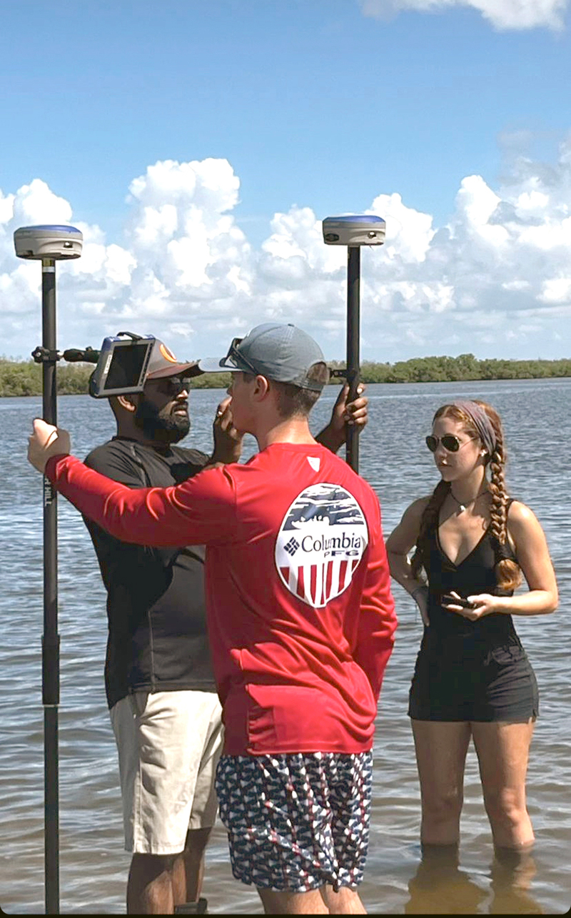 Three people stand in shallow water near a large body of water under a partly cloudy sky. Two men hold tall poles with GPS devices, and a woman stands nearby observing. They appear to be conducting a survey or fieldwork.