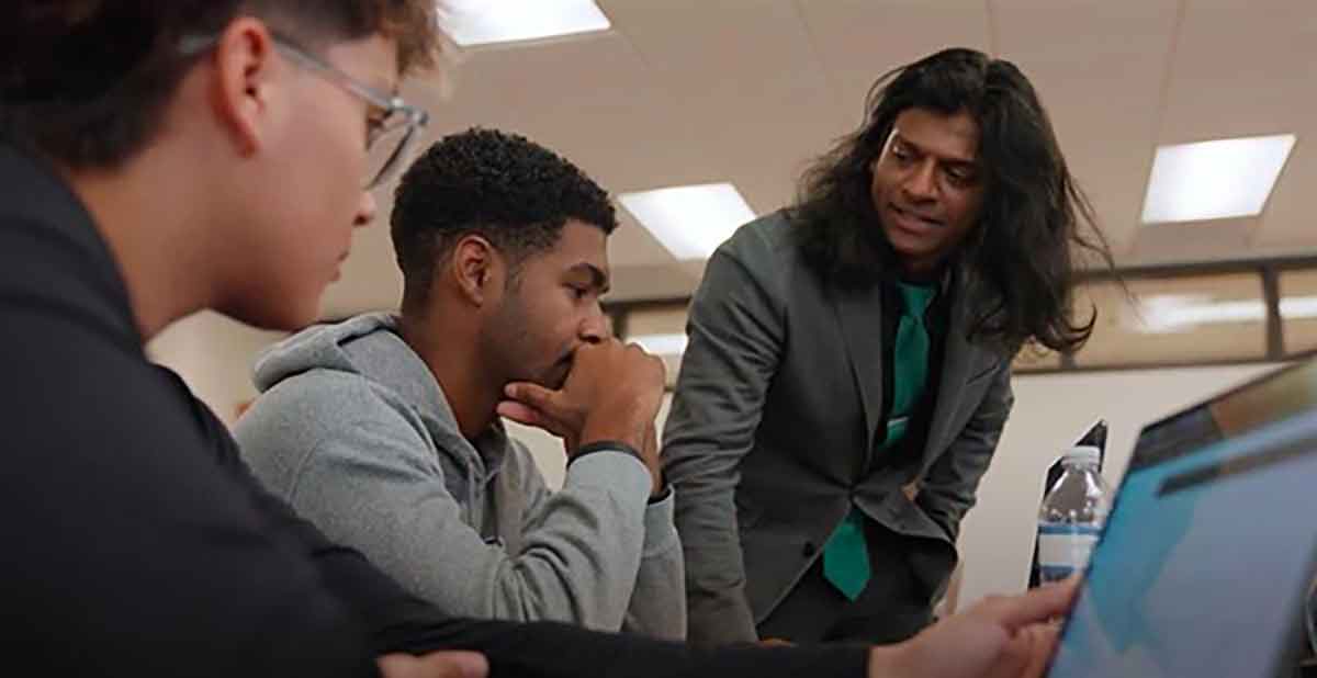A long-haired man in the background with two students in the foreground