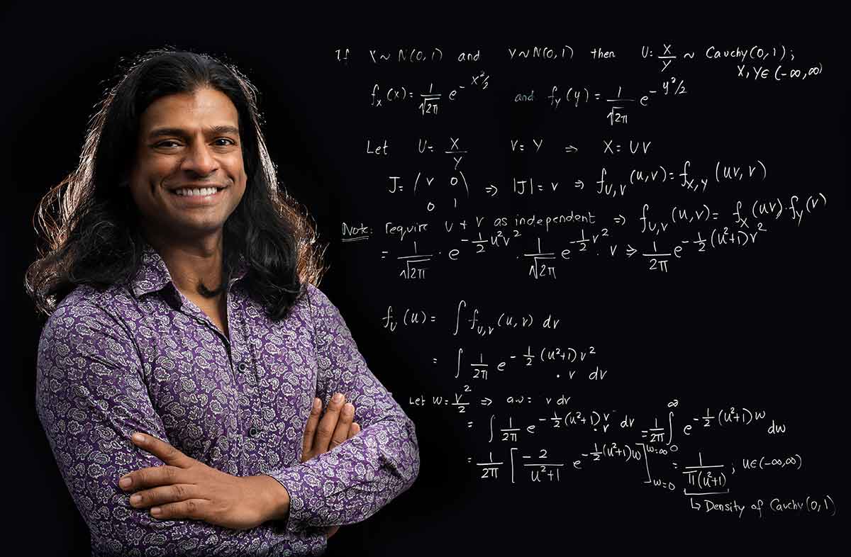A long-haired man in a purple button-down shirt stands in front of a blackboard with math writing