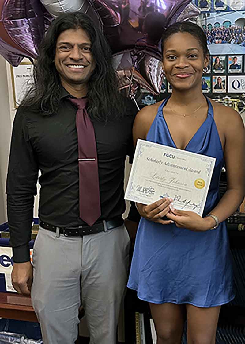 A man with long hair wears a black button-down shirt, maroon tie and grey slacks. He stands next to a woman holding a paper certificate, wearing a blue dress.