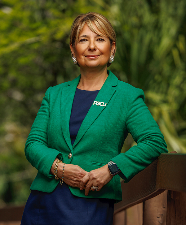 A woman stands outdoors in front of greenery, wearing a green blazer with an 'FGCU' pin on the left lapel, a navy blue top, and accessories including earrings, bracelets, rings, and a smartwatch. Their arms are crossed and resting on a wooden railing.