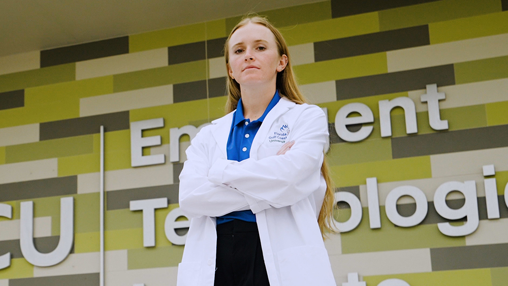 A woman with long hair, wearing a white lab coat and a blue shirt underneath, stands with arms crossed in front of a building. The building has a patterned wall with alternating shades of green and gray rectangles.