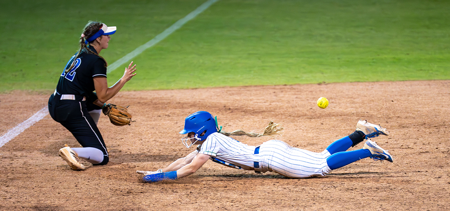A softball player in a blue helmet and striped FGCU uniform slides headfirst toward a base with outstretched arms, while a player in a black uniform kneels nearby, glove ready. A yellow baseball is mid-air between them on a dirt field with green grass in the background.