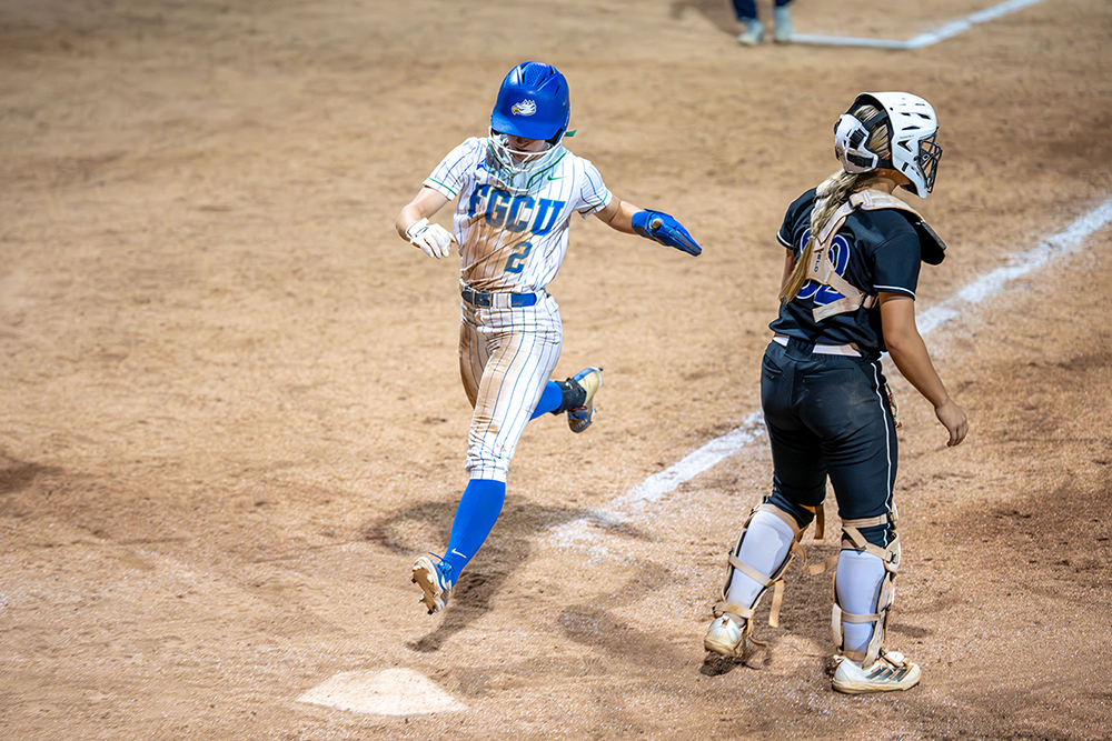 A softball player wearing a blue helmet and white FGCU uniform runs toward home plate as a catcher in black and white gear watches. The player's left foot is about to touch the base on a dirt field with chalk lines.