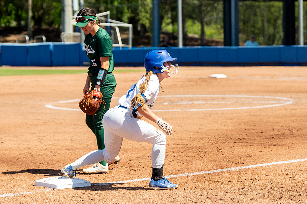 A softball player in a blue helmet and white FGCU uniform crouches at first base, ready to run, while a player in a green uniform stands behind the base with a glove.