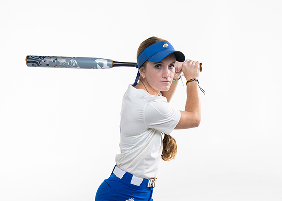 A person in a softball uniform is holding a bat in a swinging position. The person is wearing blue pants, white socks, blue shoes, and a white shirt with text and graphics.