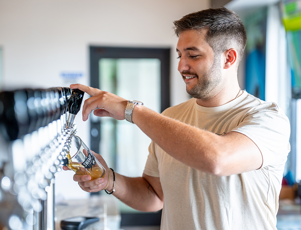 A man pouring beer from a tap into a glass. They are wearing a light-colored t-shirt and a wristwatch on their left wrist. The background features an indoor setting.