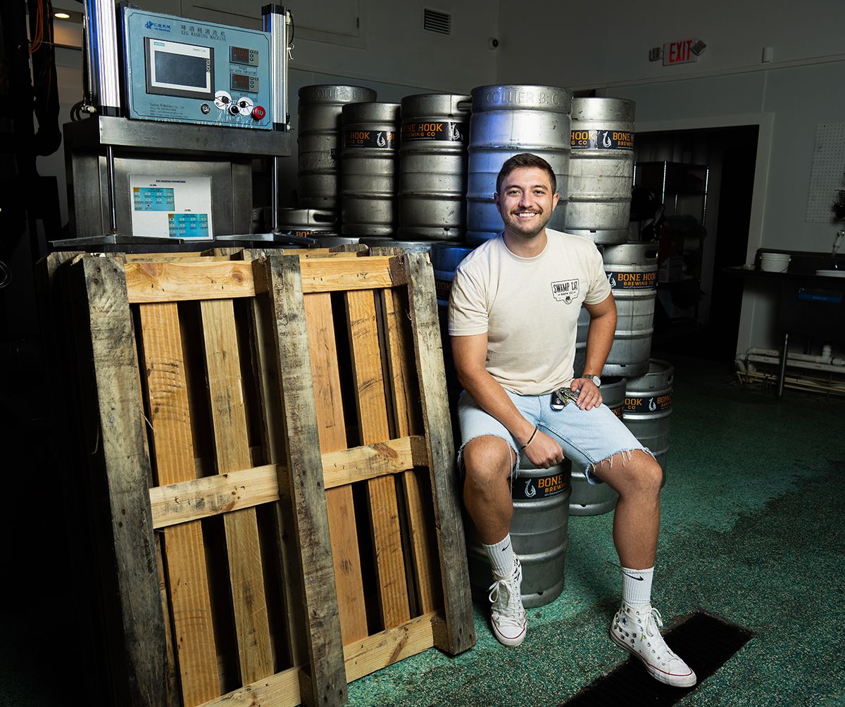 A man sitting on a stack of metal beer kegs in a brewery. They are wearing a light-colored t-shirt, light blue shorts, white socks, and white sneakers. To the left, two wooden pallets lean against each other. Behind them are multiple stacks of beer kegs labeled 'Bone Hook.' Brewing equipment and an exit sign are visible in the background.