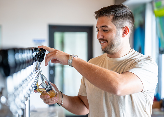 A man pouring beer from a tap into a glass. They are wearing a light-colored t-shirt and a wristwatch on their left wrist. The background features an indoor setting.