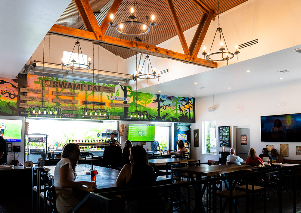 The image shows the interior of a bar or restaurant with high ceilings and wooden beams. Several tables and chairs are arranged throughout the space, with a few people seated. A colorful mural on the back wall prominently displays the words 'SWAMP CAT' along with various drink options listed below. Multiple chandeliers hang from the ceiling, and several TV screens on the walls show different programs.
