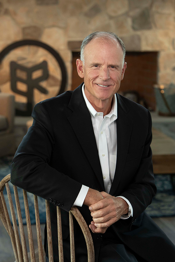 A man wearing a black suit and white dress shirt sits on a wooden chair in front of a stone wall with a fireplace. Behind him is a large circular emblem with an abstract design. The setting appears to be an indoor space, such as a living room or office