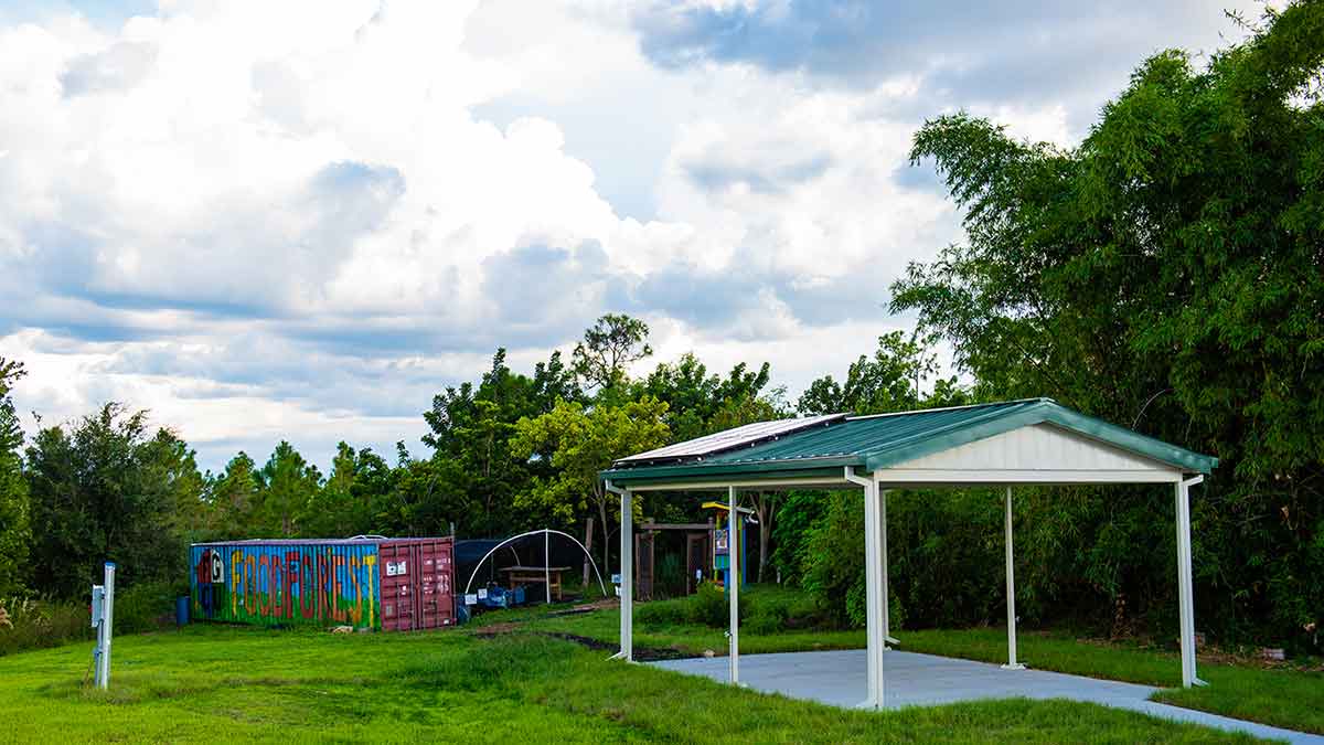A small outdoor pavilion featuring a green metal roof and white pillars on the right, set on a concrete slab, surrounded by green grass and forest in the background. On the left, a colorful shipping container displays graffiti art that reads Food Forest. Trees and bushes are visible behind the container, with a partly cloudy sky in the background