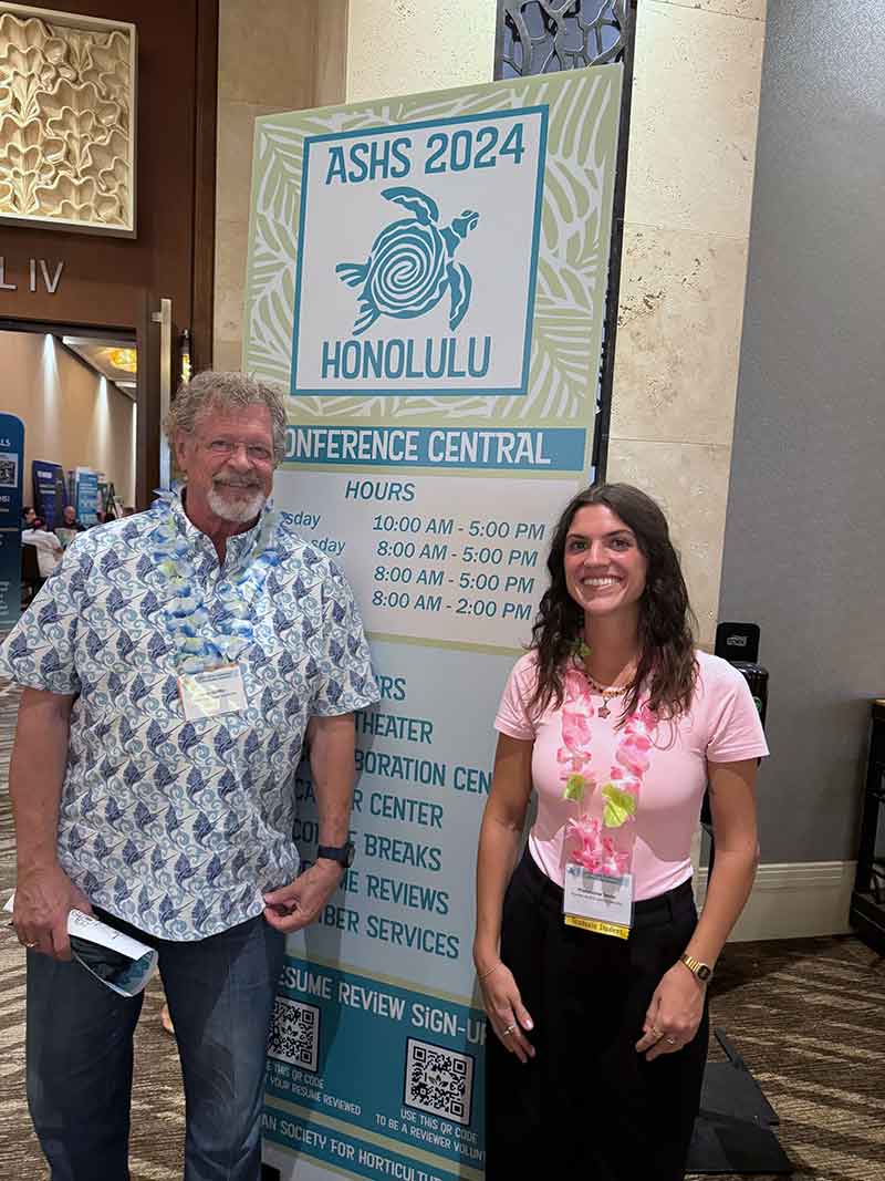 An older man and a younger brunette stand in front of a sign that reads, ASHS 2024 Honolulu Conference Central