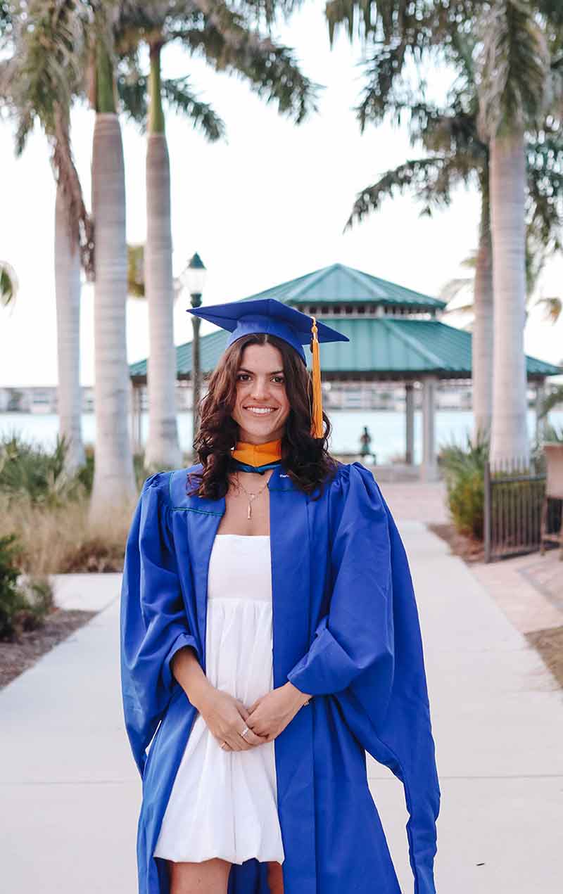 A smiling brunette, wearing a blue graduation cap and gown and white dress, stands on a paved walkway in front of a green roofed chickee hut in front of a waterfront