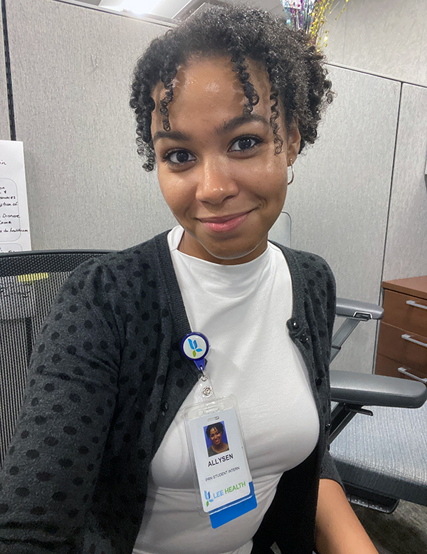 A person is sitting in an office chair, wearing a white shirt and a black polka dot cardigan. A name badge clipped to their shirt reads 'ALLYSON' with the title 'ASSOCIATE EDITOR' and the 'LEE HEALTH' logo. The background includes a grey cubicle wall, part of a wooden desk with drawers, and colorful decorations on top of the cubicle wall.