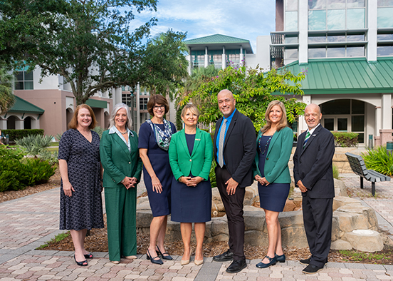 A group of seven adults, dressed in business attire, stand outdoors on a paved area with a building and trees in the background. The individuals are arranged in a line, facing the camera. The building behind them has green roofs and large windows.