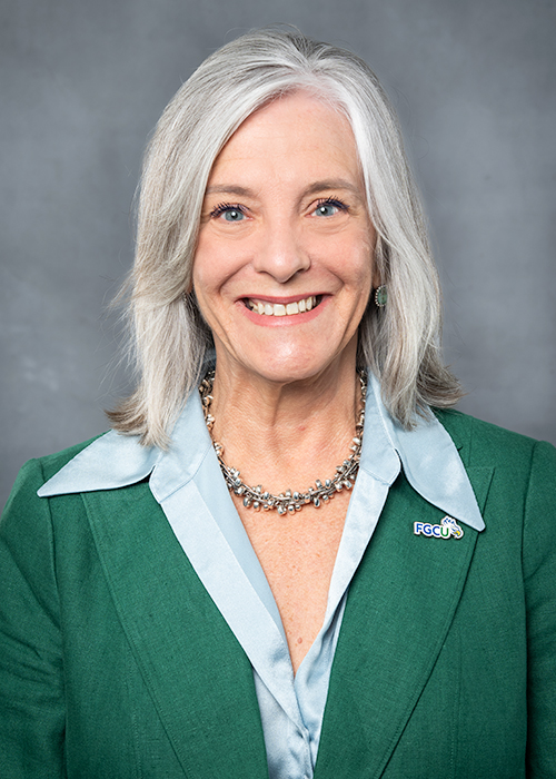 A person with shoulder-length gray hair is wearing a green blazer over a light blue collared shirt. The blazer has an FGCU (Florida Gulf Coast University) pin on the left lapel. The background is a plain gray backdrop.