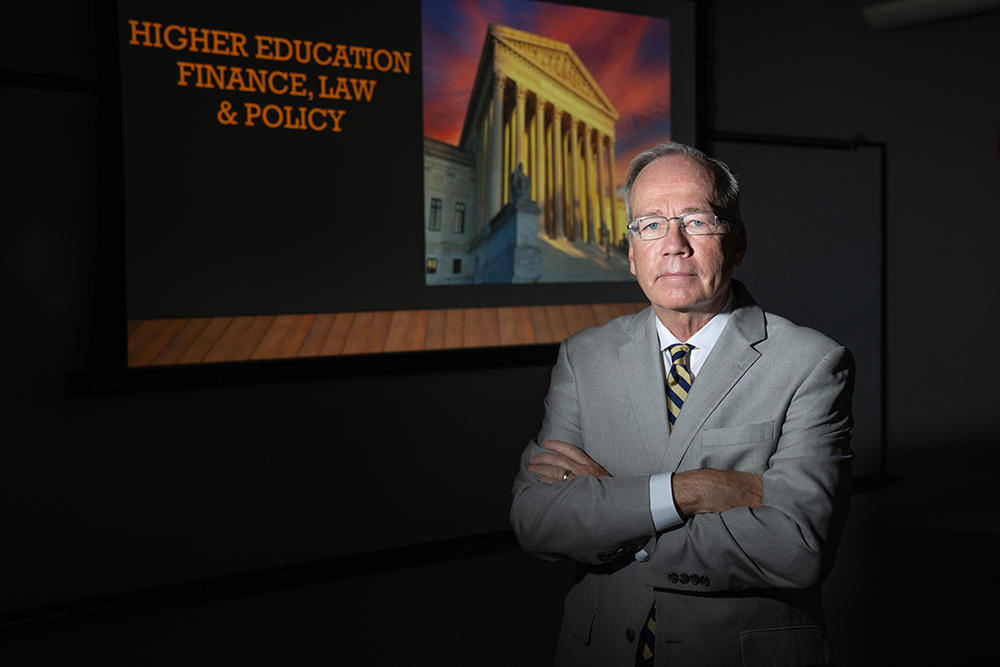 A man in a gray suit stands with arms crossed in front of a projection screen displaying the U.S. Supreme Court building and the words “HIGHER EDUCATION FINANCE, LAW & POLICY.”