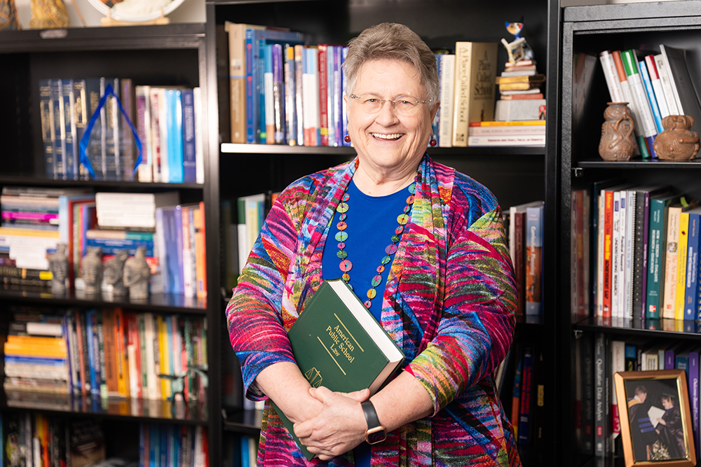 A woman wearing glasses, a colorful patterned jacket, and a beaded necklace holds a green book titled “The History of Mathematics” in front of a bookshelf filled with books, framed photos, and owl figurines.