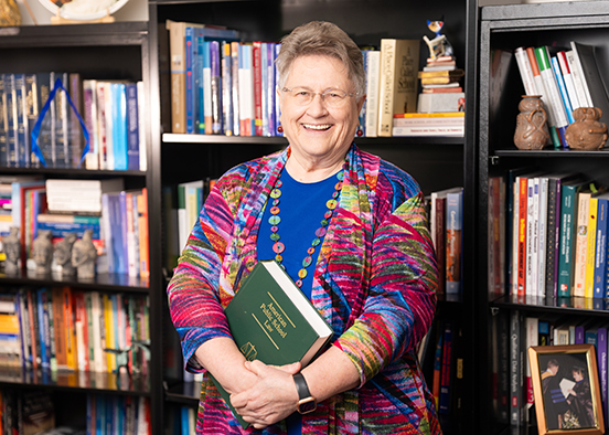 A woman wearing glasses, a colorful patterned jacket, and a beaded necklace holds a green book titled “The History of Mathematics” in front of a bookshelf filled with books, framed photos, and owl figurines.
