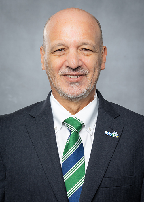 A person wearing a dark suit jacket, white dress shirt, and a green and blue striped tie. There is an FGCU (Florida Gulf Coast University) pin on the left lapel of the suit jacket. The background is plain gray.