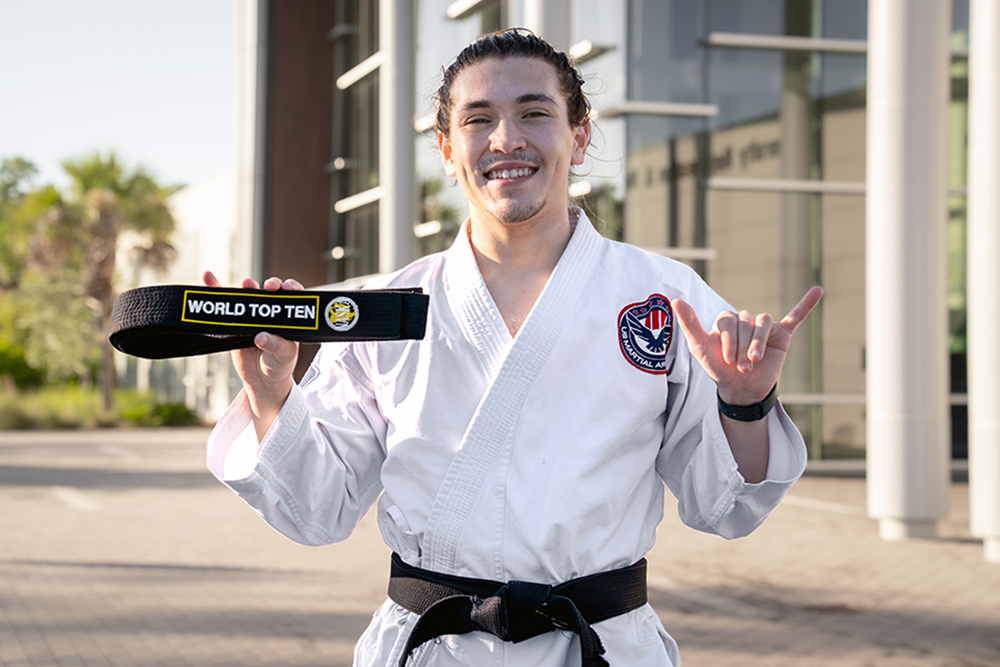 A person in a white martial arts uniform with a black belt stands outside in front of a modern building. They hold up a black belt labeled 'WORLD TOP TEN' with an emblem, and make a hand gesture with the right hand, extending the thumb and pinky. A patch is visible on the left chest of the uniform.