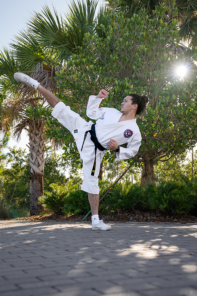A person in a white martial arts uniform with a black belt performs a high kick outdoors on a gray brick path, surrounded by lush green trees and palm plants with sunlight filtering through.