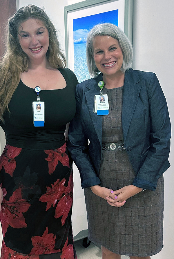 Two individuals are standing side by side in an indoor setting. The individual on the left has long, wavy hair and is wearing a black top with a red floral skirt. The individual on the right has short, gray hair and is wearing a gray dress with a dark blazer. Both are wearing identification badges around their necks. In the background, a framed picture of an ocean scene hangs on the wall.