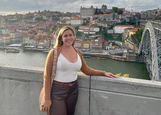 Woman standing in front of a concrete railing overlooking a scenic city with red-tiled rooftops, a river, boats, and a large bridge under a partly cloudy sky.