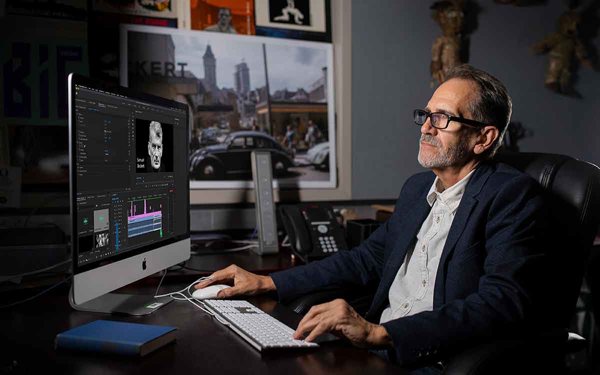 A photo of a man wearing glasses, a white button-down shirt and black blazer, seated at a desk working at a computer