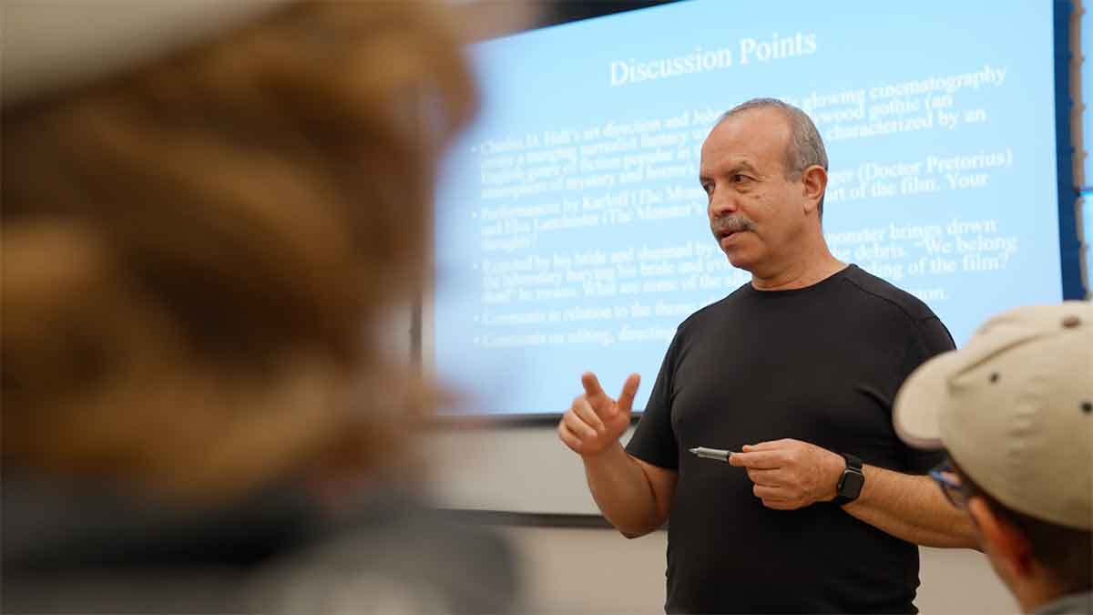 Blurry image of a student in the foreground and a professor in a black t-shirt in front of a blue PowerPoint presentation on a screen behind him