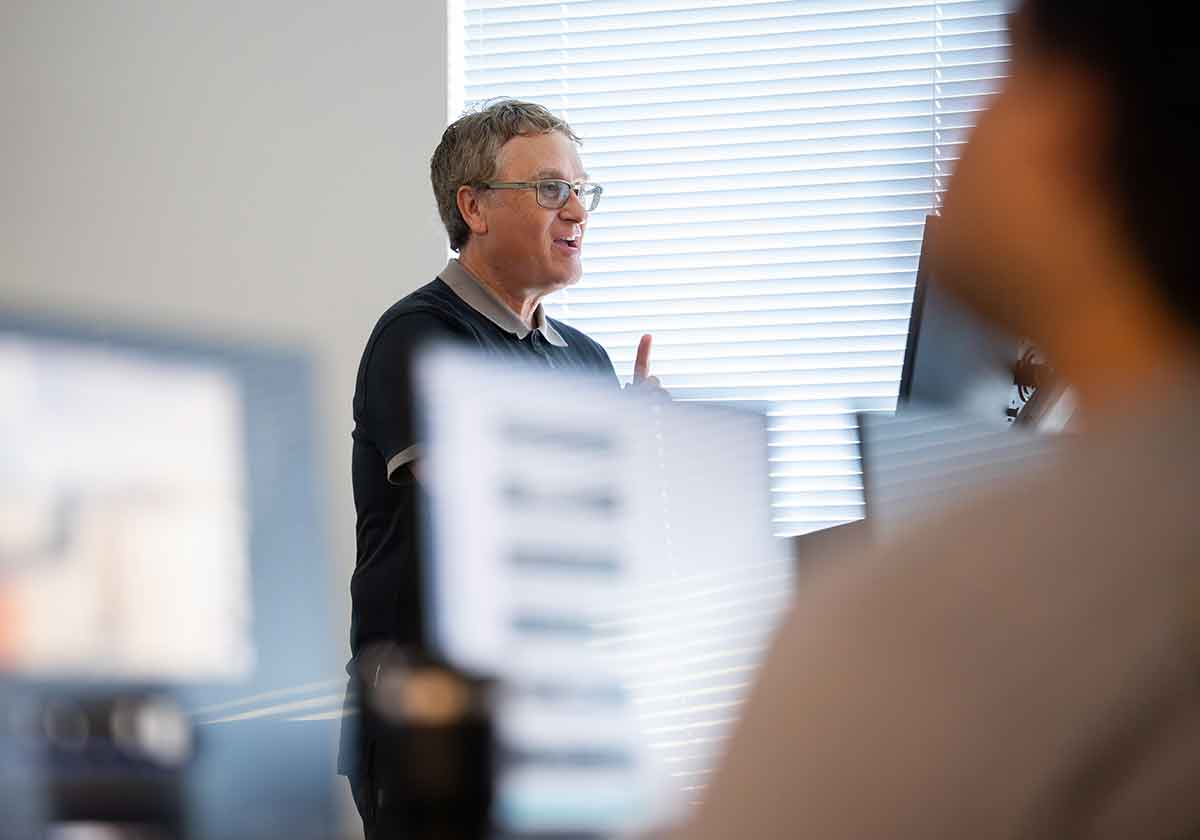 Instructor standing near a window with blinds, speaking to a class with computer monitors in the foreground