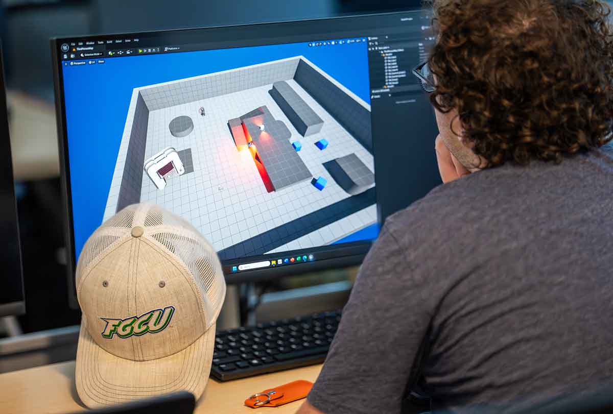 Person working on a computer showing a 3D game design interface with a gray grid and colored objects
