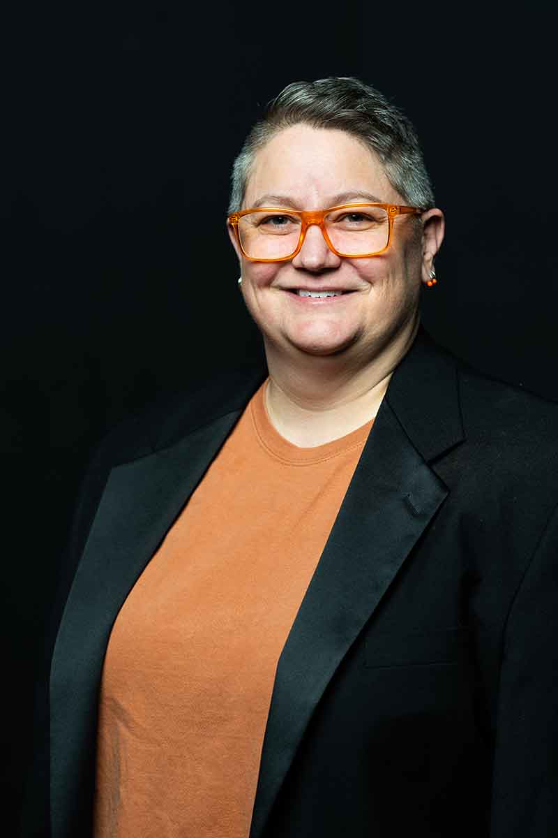 A headshot of a woman with short hair, red-rimmed glasses, an orange shirt and black tuxedo blazer