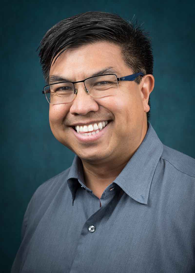 A headshot of a smiling man with dark hair and glasses in a grey, button-down shirt