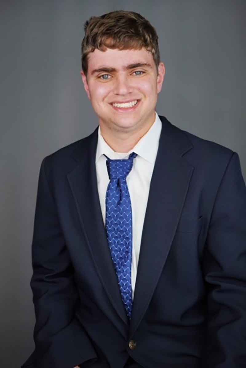 A head shot of a student in a black blazer, white button-down dress shirt and blue tie